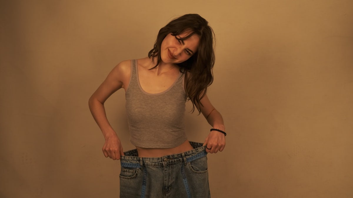 A woman smiles while holding out the waistband of oversized jeans to show her weight loss success.